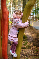 Cute happy little girl in winter faux fur ear muffs plays outdoors at wooden playground on warm sunny autumn day - 502481866