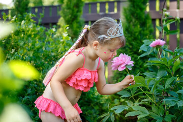 Adorable cute little caucasian four year old kid girl wearing swimming suit and princess tiara, holds in hands, smell and enjoying a pink peony flower in full bloom on the background of nature - 502481871