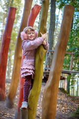 Cute happy little girl in winter faux fur ear muffs plays outdoors at wooden playground on warm sunny autumn day - 502481881