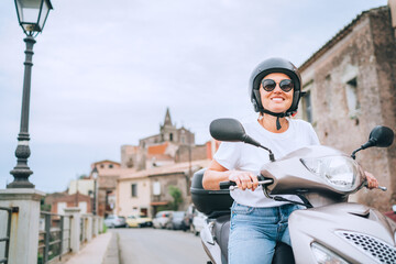 Fototapeta na wymiar Cheerfully smiling woman in helmet and sunglasses fast riding the moto scooter on the Sicilian old town streets in the Forza d'Agro. Happy Italian vacation and transportation concept.