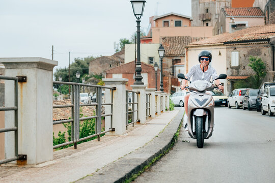 Fototapeta Cheerfully smiling man in helmet and sunglasses fast riding the moto scooter on the Sicilian old town streets in the Forza d'Agro. Happy Italian vacation and transportation concept.