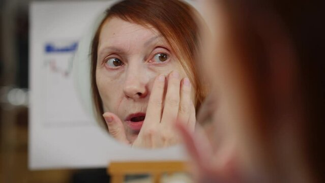 Close-up reflection in mirror of anxious mature Caucasian woman touching wrinkles. Worried lady sitting in office checking face skin. Aging and femininity concept