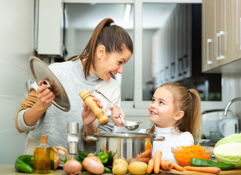 Little daughter helping cooking soup and mother add pepper to pan