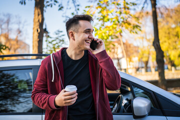 Young caucasian man standing by his car in autumn day or evening holding a cup of takeaway coffee looking to the side while waiting on the street in town copy space making a phone call talking happy