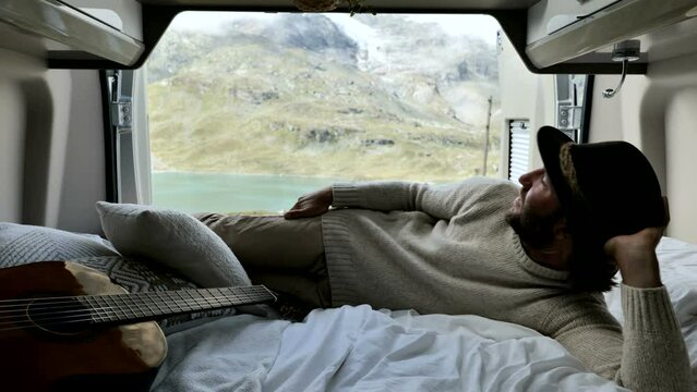 Man Traveller Lying Down In Is Camper Van Bed With Stunning Mountain Lake View. Musician In The Back Of His Van Travelling In Summer