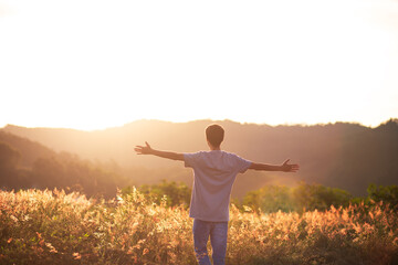 Young man sitting on the top of the mountain with his arms raised celebrating or thanking, with the sunset in the background. Concept of mental well-being, happiness, success and freedom.