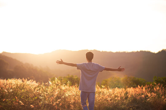 Young man sitting on the top of the mountain with his arms raised celebrating or thanking, with the sunset in the background. Concept of mental well-being, happiness, success and freedom.