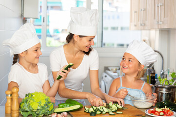 Woman with two kids in ches hats cooking together cutting and tasting vegetables for salad at home kitchen