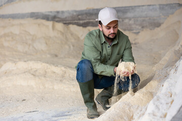 Male farmer squatting at heap of corn flour and holding bunch of it in hands.