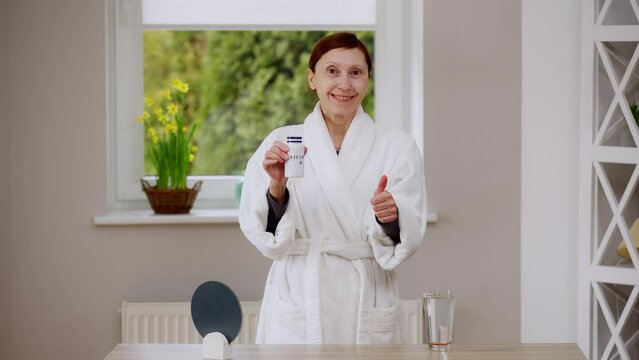 Confident happy mature woman advertising Vitamin B complex gesturing thumb up smiling looking at camera. Front view portrait of Caucasian retiree posing at home indoors with supplement