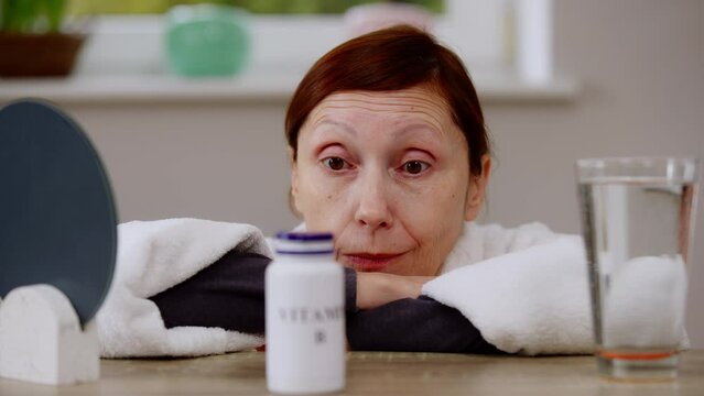 Close-up thoughtful mature Caucasian woman sitting at table with vitamins bottle. Front view portrait of female retire thinking in the morning at home. Pharmacy and health care concept