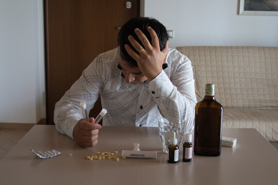 Worried man sitting and leaning on the table at home with anxiolytics and antidepressants in front while holding a test tube in anus. Side effects of psychiatric drugs on male ejaculation