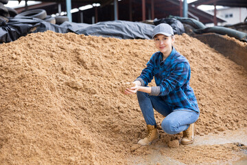 Positive woman farmer crouched down near big pile of beer bagasse, natural cattle feed, checking quality of forage - obrazy, fototapety, plakaty