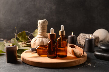 Wooden board with bottles of essential oil, sea salt, massage bag and burning candles on dark background - obrazy, fototapety, plakaty