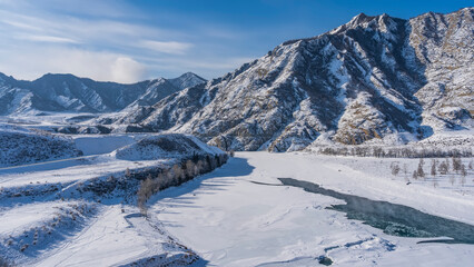 A frozen river in a snowy valley. Steam rises from the partially thawed turquoise water. Bare trees on the banks. A picturesque mountain range with steep slopes against a blue sky. Altai. Katun