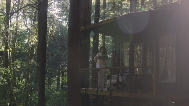 Young woman reading a book and drinking hot tea on a porch of a cozy cabin in the woods. Log cabin in the middle of woods sunny wooden bright mountain cabin rustic house architecture. 