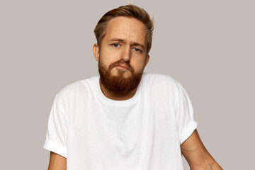 Obraz premium Doubtful young man shrugging with pursed screwed lips finding no excuse for being late at work, dressed casually in white t-shirt with blank copy space for your promotional content, isolated on gray