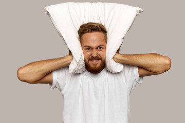 Mad furious man of 30s pressing pillow tightly to his ears, clenching teeth with anger, feeling...