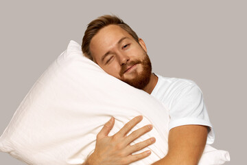 Horizontal image of smiling relaxed red-haired Caucasian man in white home clothes hugging pillow,...