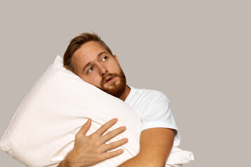 Portrait of sad attractive redhead male hipster with mild insomnia, hugging pillow looking up counting sheep in mind to fall asleep quickly, isolated over gray studio wall background