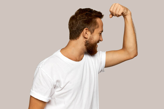 Happy cheerful bearded guy in white sport clothes laughing looking at his biceps, feeling ashamed of wick muscles, standing half-turned to camera against gray studio wall. Out of shape