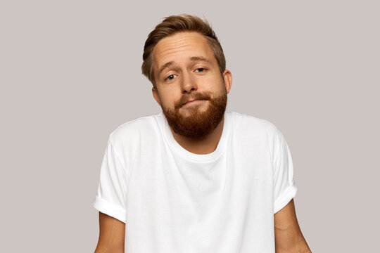 Young man with hipster trendy haircut and bearded chin shrugging over gray wall at studio, pursing lips, having no words to explain his behavior, looking at camera with uncertain smile