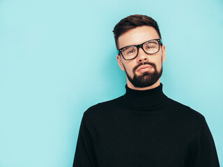 Obraz premium Handsome smiling model. Sexy stylish man dressed in black turtleneck sweater and jeans. Fashion hipster male posing near blue wall in studio. Isolated. In spectacles or eyeglasses