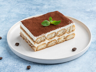 Tiramisu cake decorated with cocoa powder and fresh green mint leaf. Close up food. Blue background. Traditional italian dessert. Piece of cake on white ceramic plate.