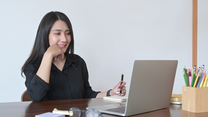 Happy young asian woman looking at laptop computer screen and making notes on notebook.