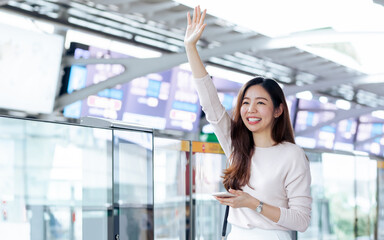 Asian working woman using mobile phoen and wave her hand to greeting her friends at train or railway station. Transportation, New Normal and Lifestyle Concept.