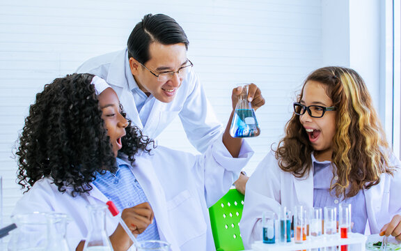 Asian handsome male teacher wearing eyeglasses and white gown uniform, teaching science and experiment to African black and Caucasian white female students in classroom at school. Education Concept.