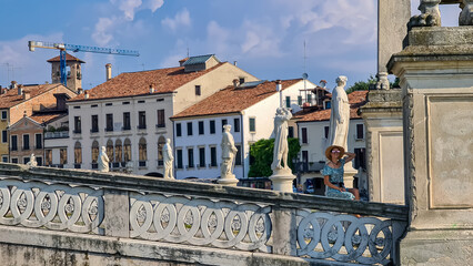 Obraz premium Woman in a dress and hat sitting on bridge of Prato della Valle, square in the city of Padua, Veneto, Italy, Europe. Isola Memmia surrounded by canal bordered by two rings of statues. Reflection water