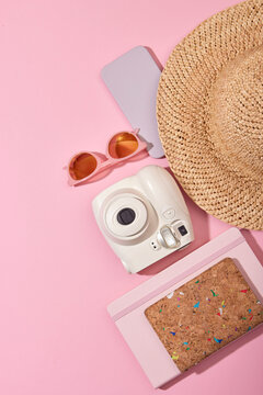 From above of instant photo camera with sunglasses smartphone and straw hat placed near notebook on pink background