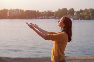 Young woman raises her hands to setting sun on shore lake, in yellow sweater enjoys life. Happy...