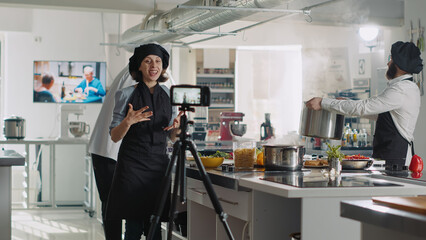 Female chef recording cooking show video on TV program, filming culinary vlog with camera in professional restaurant kitchen. Woman in uniform talking about gastronomy at online cuisine class.