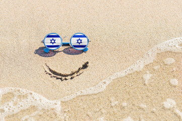 Fototapeta na wymiar A painted smile on the beach and sunglasses with the flag of Israel. The concept of a positive holiday in the resort of Israel.