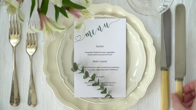 Wedding or party place setting. Hand comes into shot to add a knife to right side of frame. Flat lay footage of table arrangement. Menu card, plate, forks. knives and glasses included in shot. 