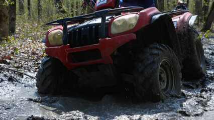 Fototapeta na wymiar Close-up of a red ATV riding through a muddy puddle in the spring forest. Extreme type of outdoor activities. Riding on a quad bike.