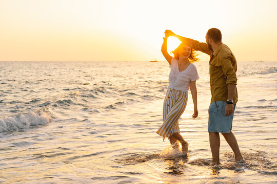 Young romantic couple dancing turning around by sea. Seascape at sunset with beautiful sky. Romantic couple on the beach at golden sunset. Lover couple having fun on beach.