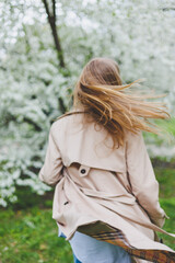 A young woman with blond hair enjoys a blooming spring garden. Travel, spring break. Fashionable style. A woman in sunglasses and a beige trench coat runs through a flowering park. Selection focus