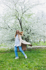 Naklejka premium A young woman with blond hair enjoys a blooming spring garden. Travel, spring break. Fashionable style. A woman in sunglasses and a beige trench coat runs through a flowering park. Selection focus