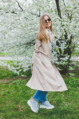 A young woman with blond hair enjoys a blooming spring garden. Travel, spring break. Fashionable style. A woman in sunglasses and a beige trench coat runs through a flowering park. Selection focus