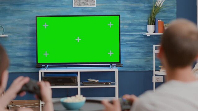 Closeup of couple holding controllers playing action console games on green screen tv sitting on couch and eating popcorn. Young gamers spending free time gaming online on chroma key mockup display.