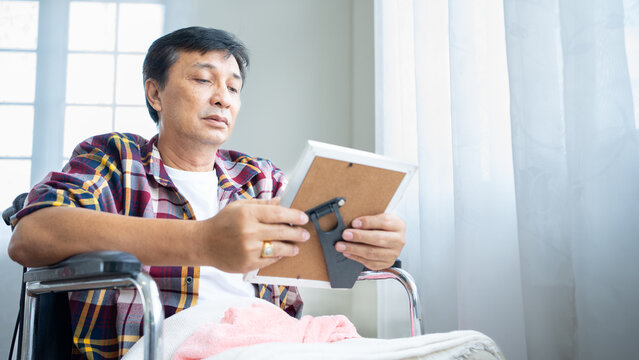 Asian Senior Male Alone On Wheelchair And Looks Family Photographs.Disability Elderly Older Mature Grandfather Feel Lonely And Sad, Missing His Relative And Wife Stay In Nursing Home After Retirement.