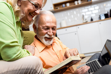 Senior couple at home spending time together, using laptop and reading book.