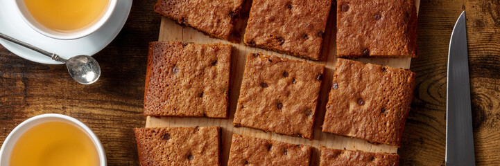 Blondie brownie dessert bar with chocolate chips, overhead flat lay panorama with tea on a dark rustic wooden background