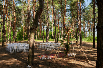 Beautiful bohemian tipi arch decoration on outdoor wedding ceremony venue in pine forest with cones. Chairs, floristic flower compositions of roses, carpet, string fairy lights. Summer rural wedding.