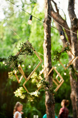 Wedding party banquet outdoors in pine forest. Dining tables, benches decorated in boho style with candles, flowers, spring fairy lights, plates, wineglasses, wooden frames. Eco organic floral decor.