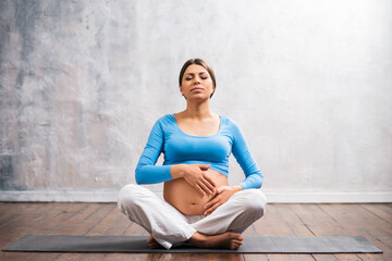 Young pregnant woman doing yoga exercises and meditating at home. Health care, mindfulness, relaxation and wellness concept.