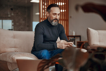 Smiling Caucasian businessman is sitting and typing on smartphone in the lobby